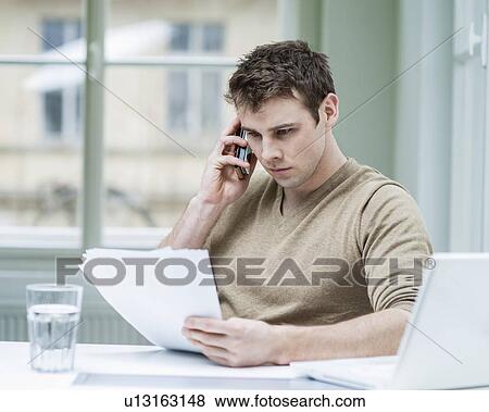 Young businessman looking at documents while using mobile phone in office View Large Photo Image Stock Photo - Young businessman looking at documents while using mobile phone in office. Fotosearch