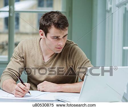 Stock Photography - Young businessman looking at laptop while writing on documents in office. Fotosearch