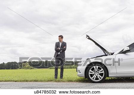 Stock Image - Young businessman standing arms crossed by broken down car at countryside. Fotosearch