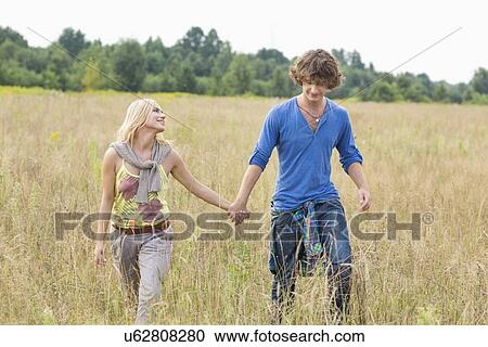 Stock Image - Young couple holding hands while walking through field. Fotosearch