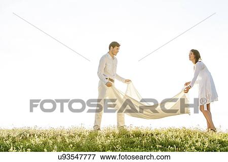 Stock Photo - Young couple spreading picnic blanket on grass during sunny day. Fotosearch