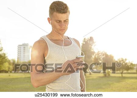 Young jogger listening to music through cell phone in park View Large Photo Image Stock Photo - Young jogger listening to music through cell phone in park. Fotosearch