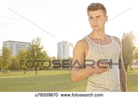 Young jogger looking away while listening music through cell phone in park View Large Photo Image Stock Image - Young jogger looking away while listening music through cell phone in park. Fotosearch
