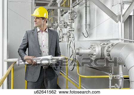 Stock Image - Young male architect holding rolled up blueprints by industrial machinery. Fotosearch