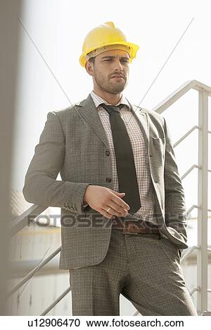 Stock Image - Young male architect in hard hat leaning on railing against clear sky. Fotosearch