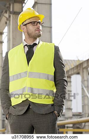 Stock Photography - Young male architect in protective workwear standing hands in pockets at construction site. Fotosearch