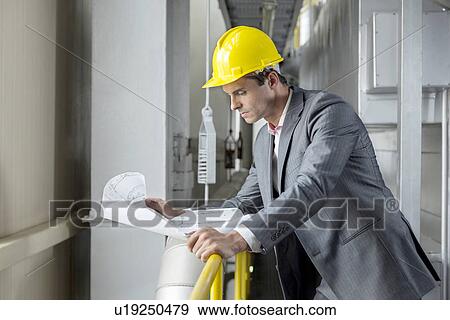 Young male architect reading blueprint while leaning on railing in industry View Large Photo Image Stock Photo - Young male architect reading blueprint while leaning on railing in industry. Fotosearch