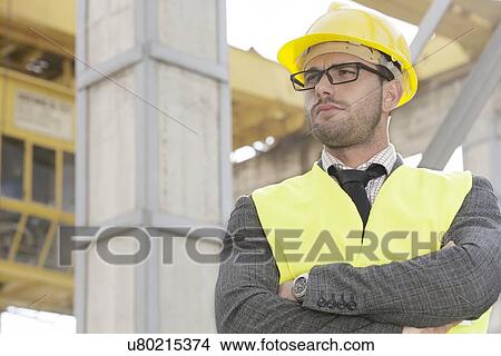Picture - Young male architect standing arms crossed at construction site. Fotosearch
