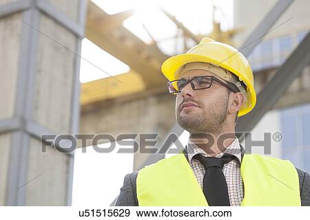 Stock Photo - Young male architect wearing hard hat looking away at construction site. Fotosearch