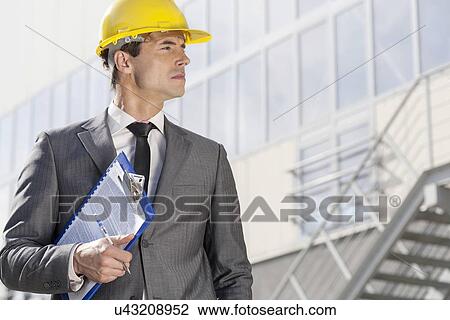 Stock Image - Young male architect with clipboard looking away outside office building. Fotosearch