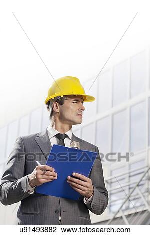 Young male architect with clipboard looking away in front of office building View Large Photo Image Stock Image - Young male architect with clipboard looking away in front of office building. Fotosearch