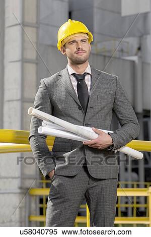 Stock Photography - Young male architect with rolled up blueprints at construction site. Fotosearch
