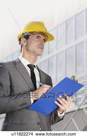 Picture - Young male architect writing on clipboard while looking away outside office building. Fotosearch