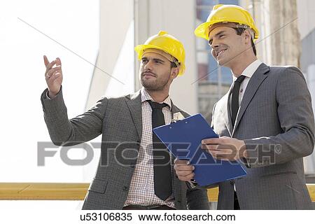 Stock Photography - Young male architects with clipboard discussing at construction site. Fotosearch