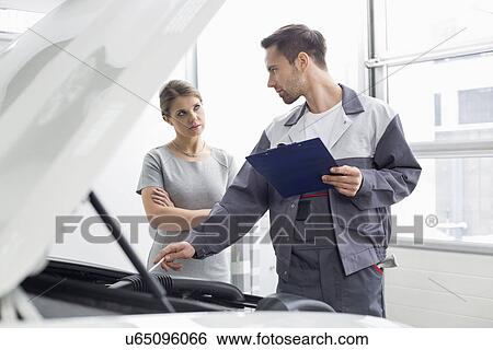 Stock Photograph - Young male engineer explaining car engine to female customer in workshop. Fotosearch