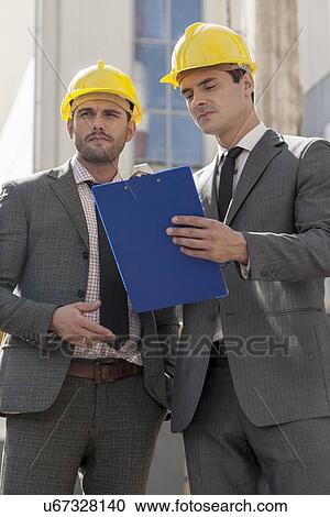 Young male engineers with clipboard discussing at construction site View Large Photo Image Stock Image - Young male engineers with clipboard discussing at construction site. Fotosearch