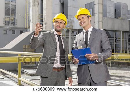 Young male engineers with clipboard discussing outside industry View Large Photo Image Stock Photo - Young male engineers with clipboard discussing outside industry. Fotosearch
