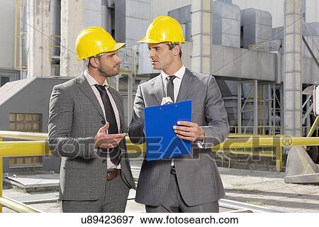 Stock Photo - Young male engineers with clipboard discussing at construction site. Fotosearch