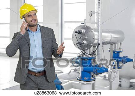 Stock Photo - Young male manager in hard hat using cell phone by machinery in industry. Fotosearch