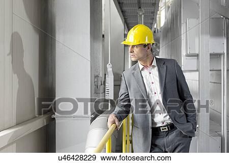 Young male manager wearing hard hat looking away in industry View Large Photo Image Stock Photo - Young male manager wearing hard hat looking away in industry. Fotosearch