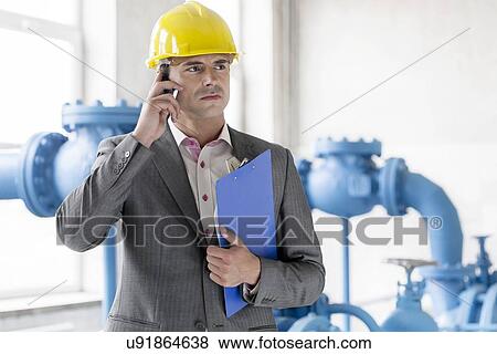 Stock Photo - Young male manager with clipboard using cell phone in industry. Fotosearch