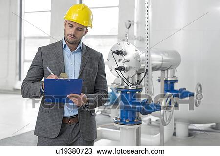 Stock Image - Young male manager writing on clipboard in industry. Fotosearch