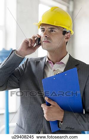 Stock Photography - Young male supervisor with clipboard using cell phone in industry. Fotosearch