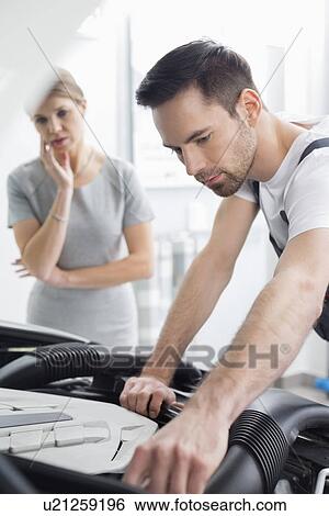 Young male technician examining car engine with worried customer in background at workshop View Large Photo Image Stock Photograph - Young male technician examining car engine with worried customer in background at workshop. Fotosearch