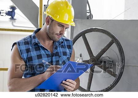 Stock Image - Young male worker writing on clipboard by large valve in industry. Fotosearch