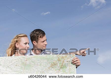 Picture - Young man holding map while woman pointing away against sky. Fotosearch
