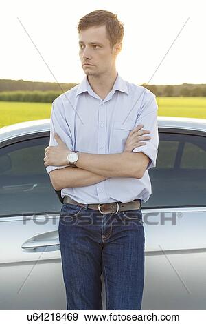Stock Photo - Young man looking away while standing by car at countryside. Fotosearch
