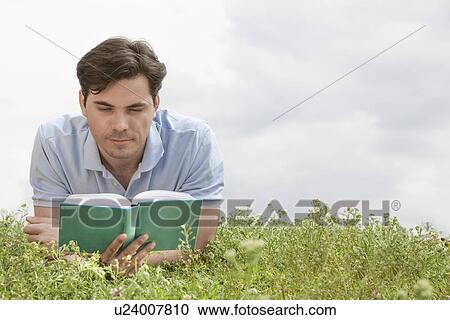 Young man reading book while lying on grass against sky View Large Photo Image Stock Image - Young man reading book while lying on grass against sky. Fotosearch