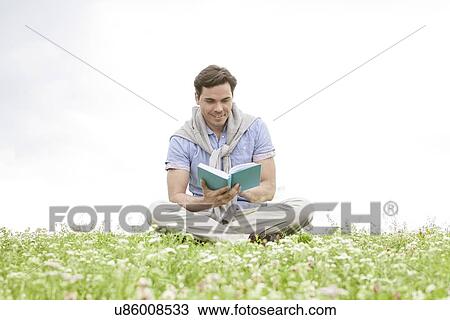 Stock Image - Young man reading book while sitting on grass against sky. Fotosearch
