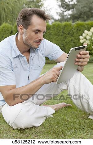 Young man using digital tablet while sitting on grass in park View Large Photo Image Stock Image - Young man using digital tablet while sitting on grass in park. Fotosearch
