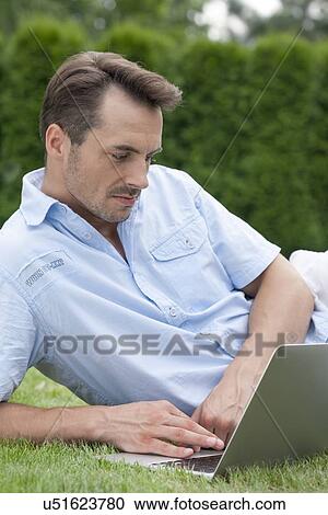 Young man using laptop while lying on grass in park View Large Photo Image Stock Image - Young man using laptop while lying on grass in park. Fotosearch