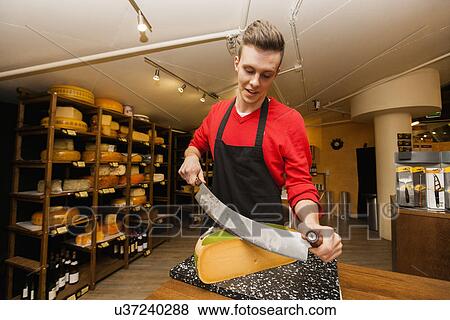Stock Photo - Young store clerk cutting cheese at counter. Fotosearch