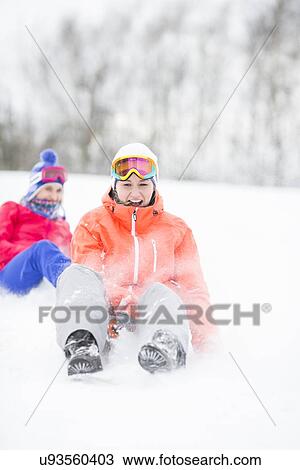 Stock Image - Young woman enjoying sled ride in snow with friend in background. Fotosearch