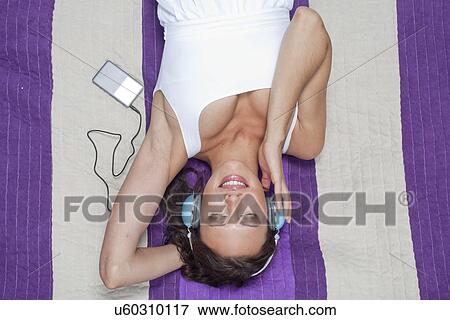 Stock Photo - Young woman listening to music through MP3 player using headphones while lying on picnic blanket. Fotosearch