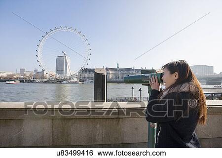 Stock Photograph - Young woman looking at London Eye through stationary viewer at London, England, UK. Fotosearch