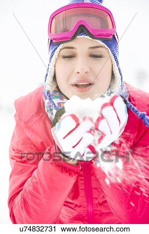 Stock Image - Young woman wearing winter coat blowing snow outdoors. Fotosearch