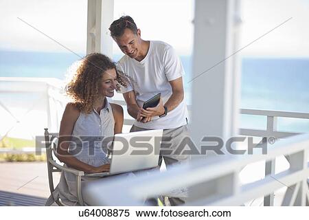 Stock Photography - Couple looking at digital tablet and laptop on beach house balcony. Fotosearch
