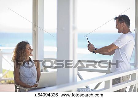 Stock Photograph - Couple taking photographs on beach house balcony. Fotosearch