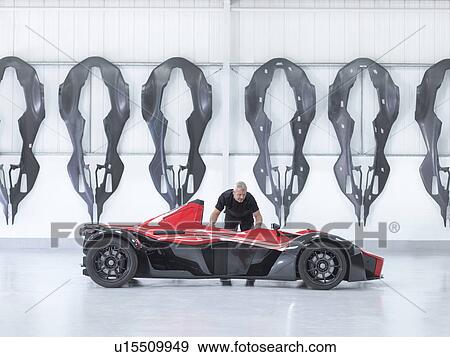 Stock Photo - Engineer inspects supercar in factory with carbon fibre car body shells hanging on wall. Fotosearch