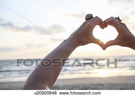 Stock Photo - Female hands making heart shape at sunset on beach. Fotosearch