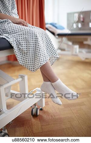 Stock Image - Girl sitting on hospital bed, wearing examination gown. Fotosearch