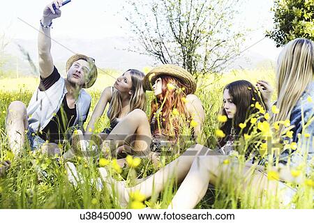 Stock Image - Group of friends sitting in grass photographing themselves. Fotosearch