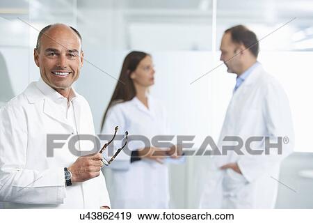 Male doctor by reflective wall, colleagues in background View Large Photo Image Stock Photo - Male doctor by reflective wall, colleagues in background. Fotosearch