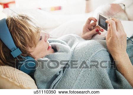 Stock Image - Mature woman lying on bed using mp3 player. Fotosearch