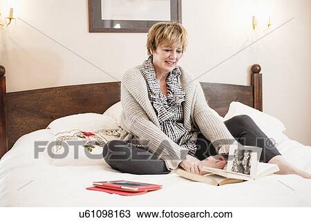 Stock Image - Mature woman sitting on bed reading book. Fotosearch