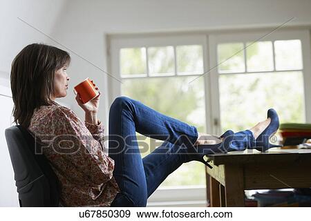 Stock Photo - Mature woman with feet on desk, drinking coffee. Fotosearch